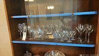 Shelf with multiple clear stemmed glasses including wine, cordial and coupe glasses, some hand-painted and etched