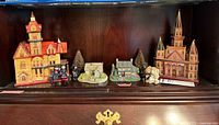 Full view of the collection on a bottom shelf, showing multiple miniature village buildings including houses, a church, human figurines, trees, and a teddy bear.