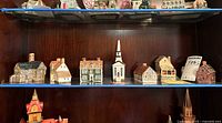 Mid-level shelf of wooden cabinet showing 11 miniature ceramic Heritage Houses and a church figurine, along with displayed boxes and pricing tag 725.