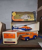 Photo shows Lionel 1956 Ford F-100 pickup truck in orange and blue with box in background and boxed Corgi Hughes 369 police helicopter on top.