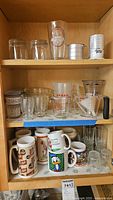 Shelf showing assorted clear glassware including measuring cups, various drinking glasses, and vintage mugs