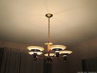 View of chandelier lit, showing the four glass shades and brass arms extending from central wood and brass column, mounted on ceiling with brass chain.