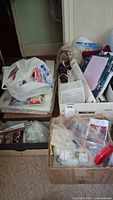 Wide view showing four boxes and bags filled with various needlepoint and craft supplies including yarn, patterns, plastic canvas, and small craft items.