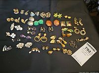 Wide view of all various non-pierced earrings lined out on black cloth. Includes multiple pairs with diverse materials like wood, enamel, rhinestones, and different shapes. Clip-ons and screw backs visible.