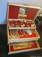 Overall view of the vintage cream jewelry box open, showing red velvet compartments containing multiple faux pearl necklaces, earrings, and a sweater clip.