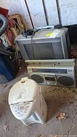 Photo of Toshiba 15-inch TV/VHS/DVD combo with Panasonic cassette boombox and Zojirushi water boiler in foreground on concrete floor with leaves and dust.