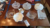Lot of seven vintage candy dishes, a creamer, and a juicer displayed on a wooden table, showing varying styles and materials including milk glass and crystal.