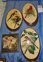 Three wood plaques laid out on blue cloth: two oval bird plaques with bark edges and one smaller framed kitten print plaque