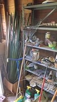 Tall green plastic coated yard stakes and wooden stakes with rakes and gloves on shelves