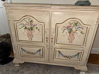 Front view of the painted wood cabinet showing two doors with floral hand-painted panels and floral garland accents on lower panels.