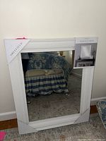 White wood decorative mirror in original packaging showing corner and part of frame and reflection of a bedroom.