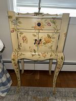 Front view of one painted floral nightstand showing top drawer, cabinet doors with black hardware, curved legs