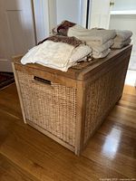 View of the wicker storage trunk with assorted stacked towels on top. Shows the trunk's natural wood frame and wicker weave.