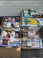 Four boxes and a plastic bin filled with assorted VHS tapes showing various titles and genres.