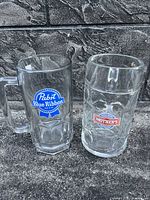 Two large beer mugs side by side with logos of Pabst Blue Ribbon and Mother's. Both are clear glass with handles and thick bases, photographed against a stone background.