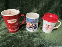 Photo showing three Tim Horton collectible mugs: one red glass mug with maple leaf and logo, one glass mug with hockey player design, and one white plastic travel mug with red lid and logo.