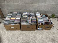 Three cardboard boxes filled with CD jewel cases, arranged side by side outdoors on concrete ground with cinder block wall background
