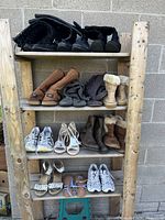 Photo showing 4 shelves of various used women's shoes including boots, sneakers, sandals and heels, arranged on wooden shelving unit outside against brick wall.