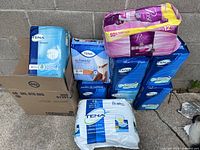 Multiple packages of women's pads including Tena Stretch, Tena Ultimate, Tena Absorbent Pads, and Poise 12 count pads shown stacked against a grey concrete wall outside.