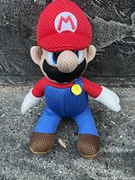 Front view of Super Mario plush toy with red hat, blue overalls and white gloves, showing detailed facial embroidery and textured fabric.