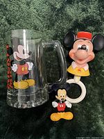 Photo showing 3 Mickey Mouse collectibles: a clear glass mug with Mickey Mouse print, a plastic keychain figure, and a toy figure head on ring handle.
