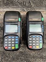 Front view of two black Pax S80 debit machines, showing displays, keypads, function buttons, and printer paper compartment with green buttons.