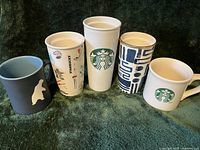 Five Starbucks mugs and tumblers displayed against a dark green background showing different designs including logos and artwork.