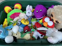 Photo showing a bin filled with various stuffed animals including recognizable characters like Barney, Elmo, and Spiderman, along with assorted animal plush toys such as a frog, sloth, bunny, bee, and more.