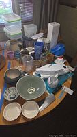 Overview of kitchen items on a round table, showing various pottery bowls, mugs in earth and blue tones, glassware, plastic storage containers with green lids, and a white handheld slicer/grater kitchen tool.