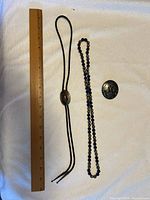 Lot display showing silver toned bolo-tie with black braided cords, dark blue beaded necklace, and oval decorative pin next to a ruler for scale.