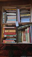 Four boxes filled with mostly hardcover books arranged upright and stacked, showing a variety of titles and authors.