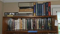 Top shelf containing striped box, white binders, rolled poster, wooden decorative box, Cleopatra book, blue cloth hardcovers, Rebecca