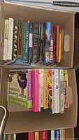 Two boxes filled with assorted cookbooks and novels, showing titles and book spines clearly.