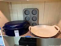 Shelf display showing dark blue enamel-coated covered roasting pan, metal muffin pan with six cups, and white enamel-coated open roasting pan