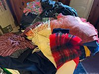 Pile of assorted scarves in red plaid, beige, pink, and other colors, partially covering clothing items including striped and solid fabrics.