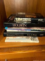 Stack of various art and architectural coffee table books on a wooden shelf, titles visible including "Imaginary Magnitude", "Holbein", "Le Corbusier", "Princeton University Bric-A-Brac 1989", and "Westminster Abbey".