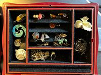 Top-down view of open box showing velvet-lined compartments filled with assorted pins, pendants, brooches, earrings and jump rings