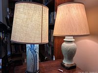 Two table lamps shown lit on a wooden surface, one with a blue and white floral pattern on a cylindrical base and one with a white textured ceramic base, both with fabric shades.