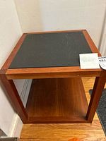 Front angle view of mid-century modern teak wood side table with a dark slate top and open teak lower shelf, placed against a wall on wooden flooring.