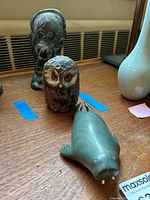 Photo showing the pottery owl figurine in front and two soapstone carvings of human figures behind it on a wooden surface.