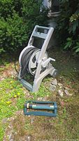 Garden hose neatly wound on gray plastic rolling reel with handle, crank, and wheels, positioned on grass and rock ground.