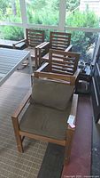 Four teak wood chairs positioned on a porch or patio, visible natural setting background, one chair with cushion installed