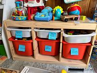 Front view of wooden toy organizer showing three large orange bins on bottom and three smaller white bins on top with blue labels.