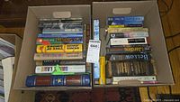 Two cardboard boxes filled with assorted hardcover and softcover books arranged vertically, showing a variety of titles and genres.