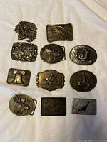 Top-down view of all 11 vintage metal belt buckles arranged on white fabric; various themes including animal, historical, western, and decorative patterns.
