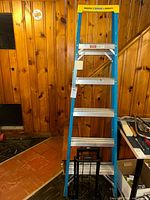 Full front view of the blue fiberglass step ladder with silver steps, standing upright indoors against a wood-paneled wall.