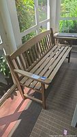 Full view of wooden porch bench placed in a sunroom or enclosed porch area with sunlight and plants visible outside.