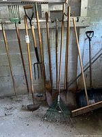 Photo shows a lineup of yard tools including rakes, forks, shovels, broom, pick axe, and spade leaning against a concrete wall.