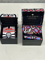 Two silk ties in individual black decorative boxes with indigenous art styled fabric on the ties and inside the lids. One tie is monochrome with red accents, the other colorful.