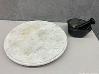 Photo showing round white marble cake stand next to heavy granite mortar and pestle on a gray surface.